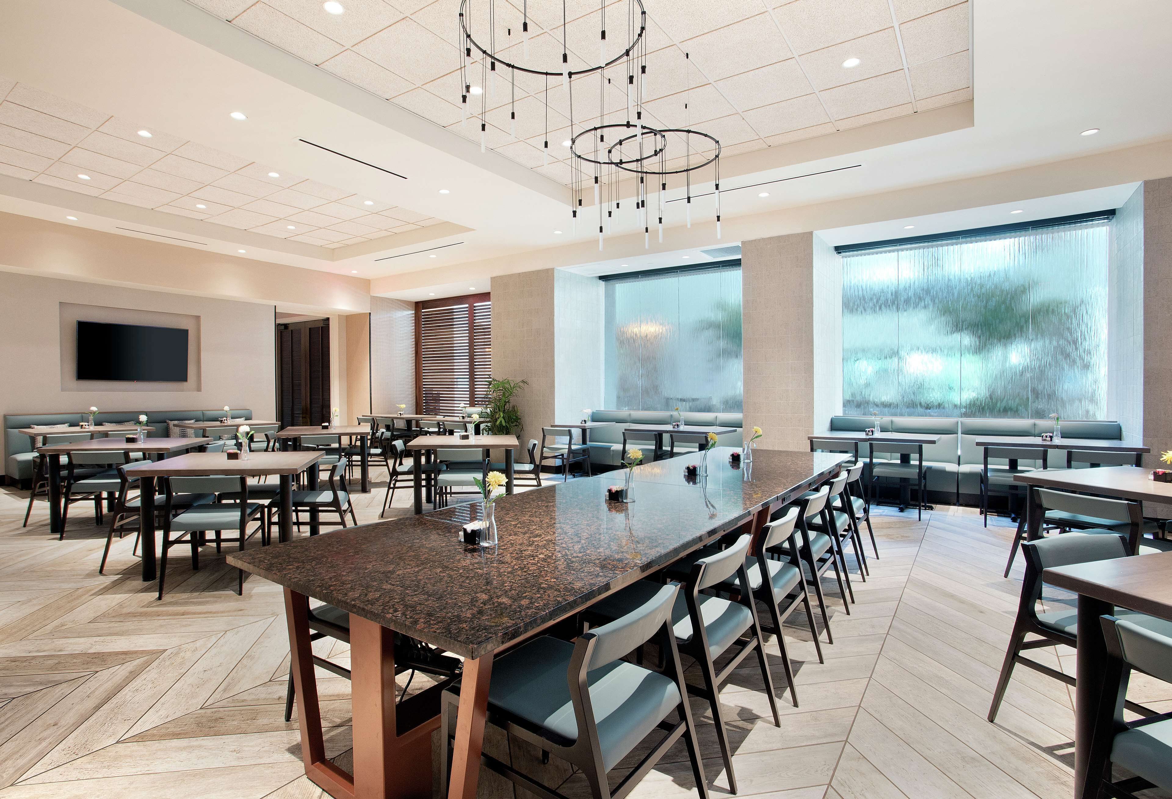Un vaste espace dédié à la salle à manger, avec de nombreuses tables et chaises, une grande table centrale et de grandes fenêtres qui laissent entrer la lumière naturelle.