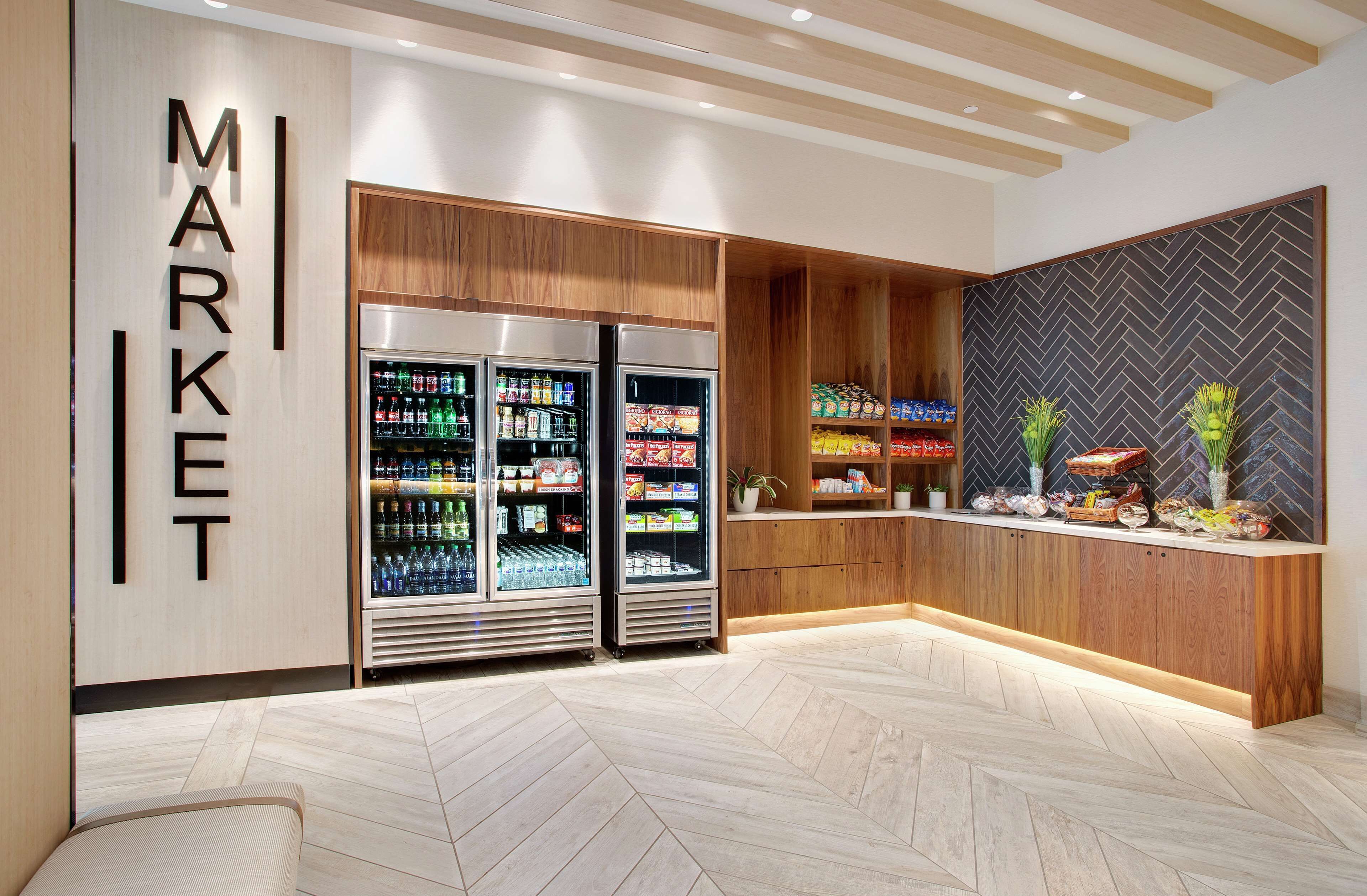 A modern market area with wooden cabinetry, a herringbone-patterned wall, and a reception desk with a display of snacks and fruits.
