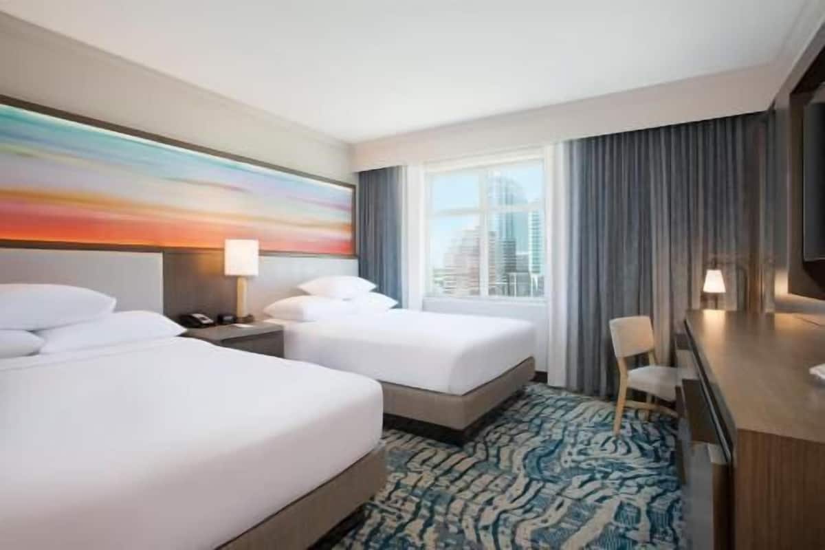 A hotel room with two beds, a desk, a chair, a large window, and a city view.