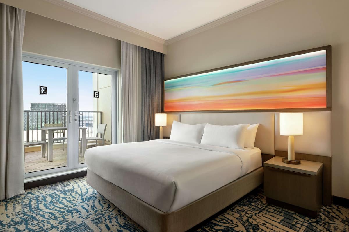 A hotel room with a large bed, two bedside lamps, a view of the outside balcony, and a colorful abstract painting on the wall.