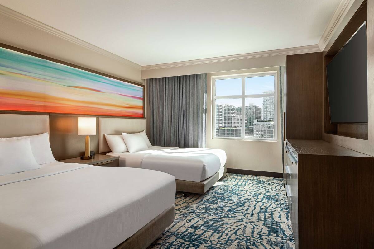 A hotel room with two beds, a large window with a city view, a flat-screen TV, and a colorful abstract painting on the wall.