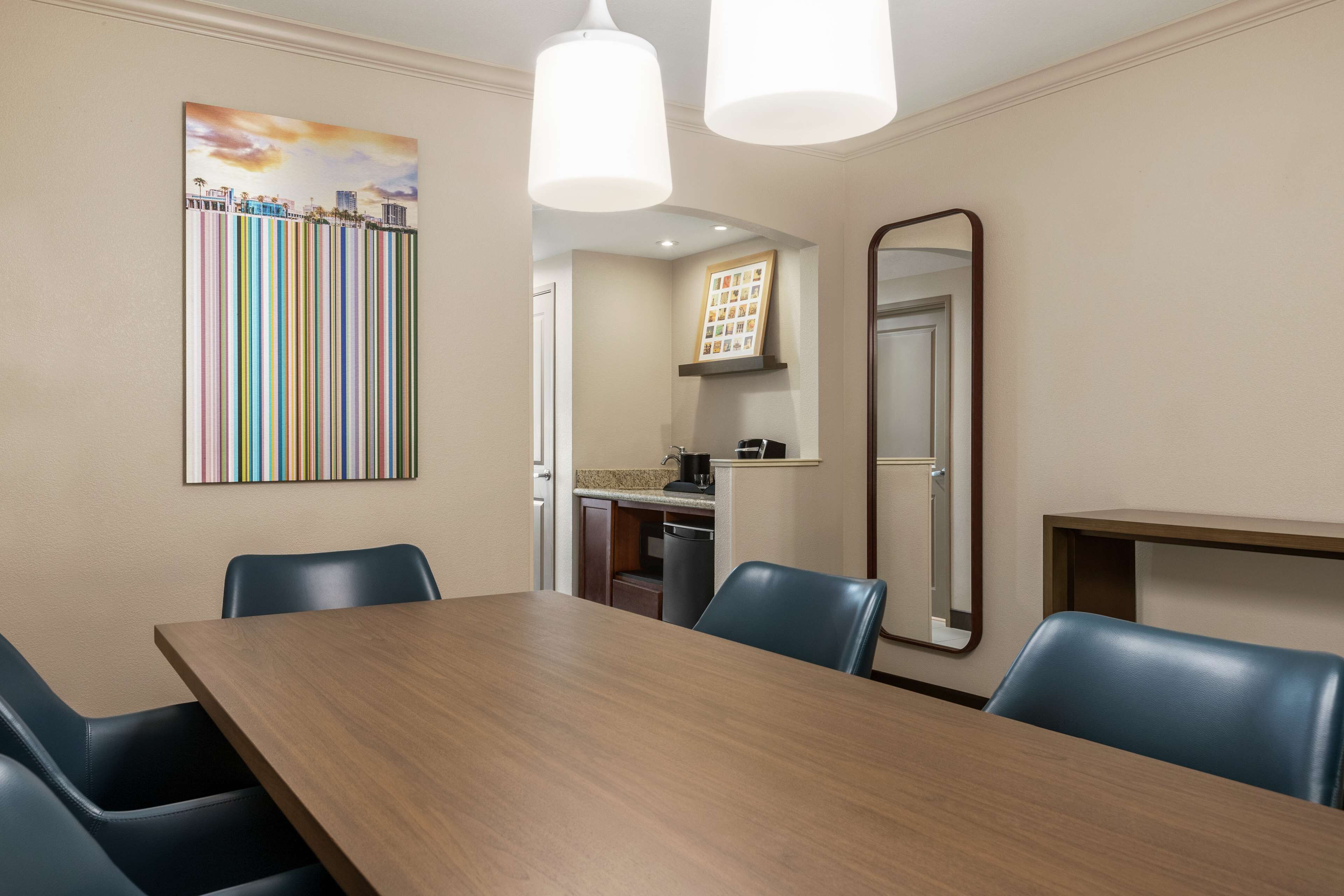 A conference room with a wooden table, blue chairs, a painting, a mirror, and a kitchenette in the background.