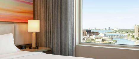 A hotel room with a bed, a nightstand, a lamp, a view of a cityscape through a window, and a colorful abstract painting on the wall.