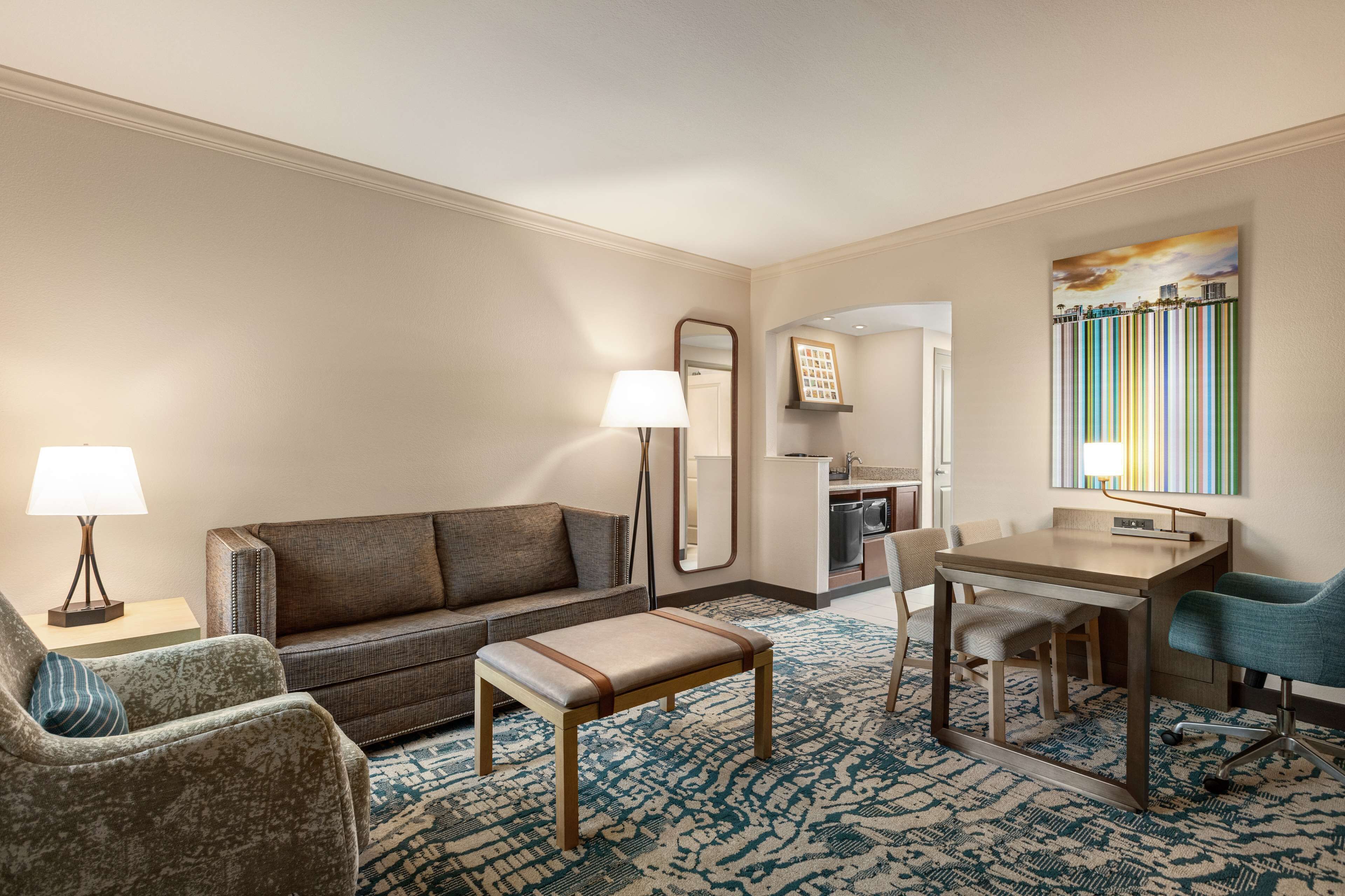 Une chambre d’hôtel comprenant un canapé, un fauteuil, un pouf, une table à manger, des chaises, une lampe, un miroir, un tableau et une kitchenette.