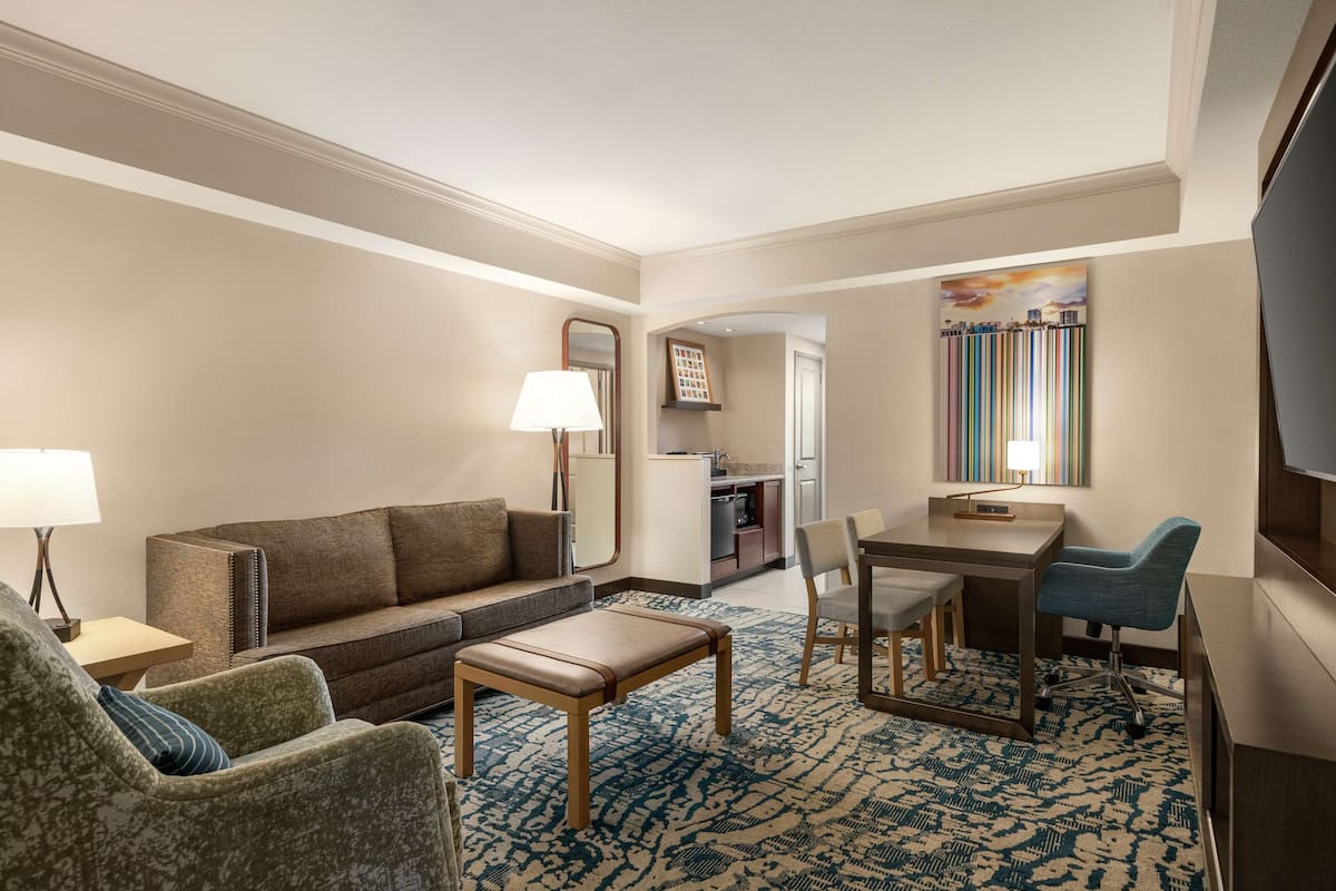A hotel room with a living area featuring a sofa, armchair, coffee table, and a dining table with chairs. There is a TV mounted on the wall and a painting above the dining area.