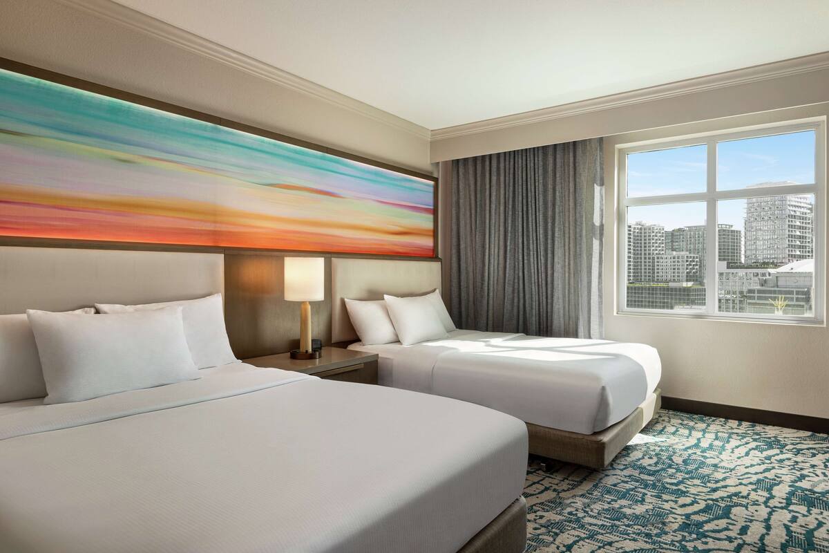 A hotel room with two beds, a large window with a city view, a modern abstract painting on the wall, and a patterned carpet.