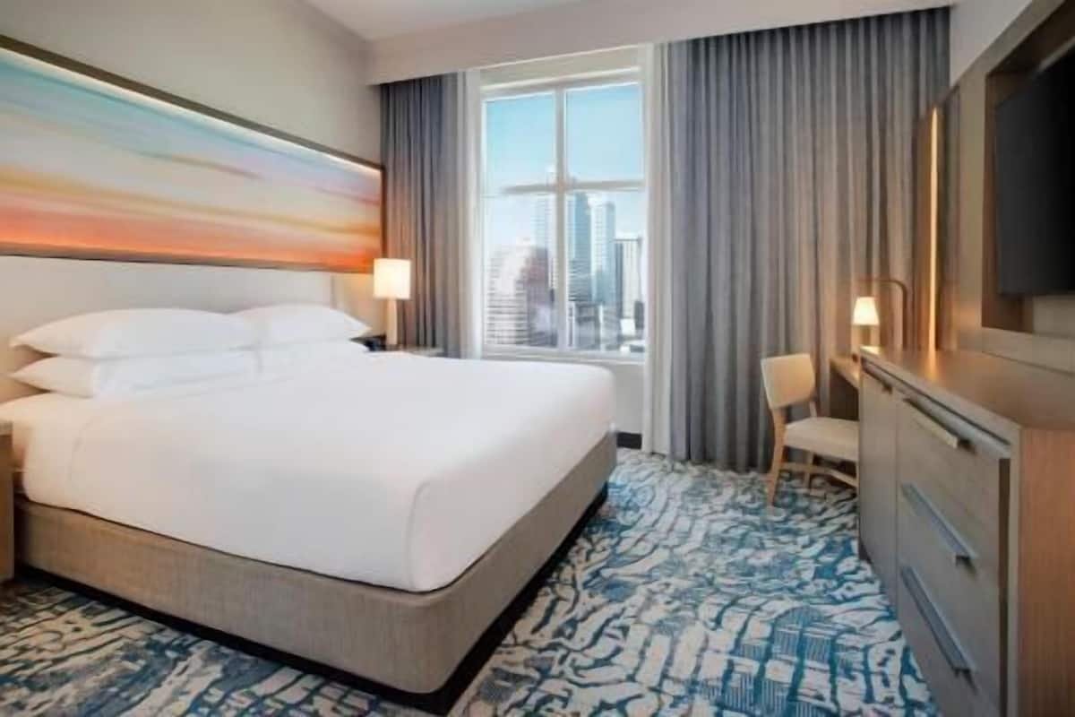 A hotel room with a large bed, a desk, a chair, a TV, and a window with a city view.