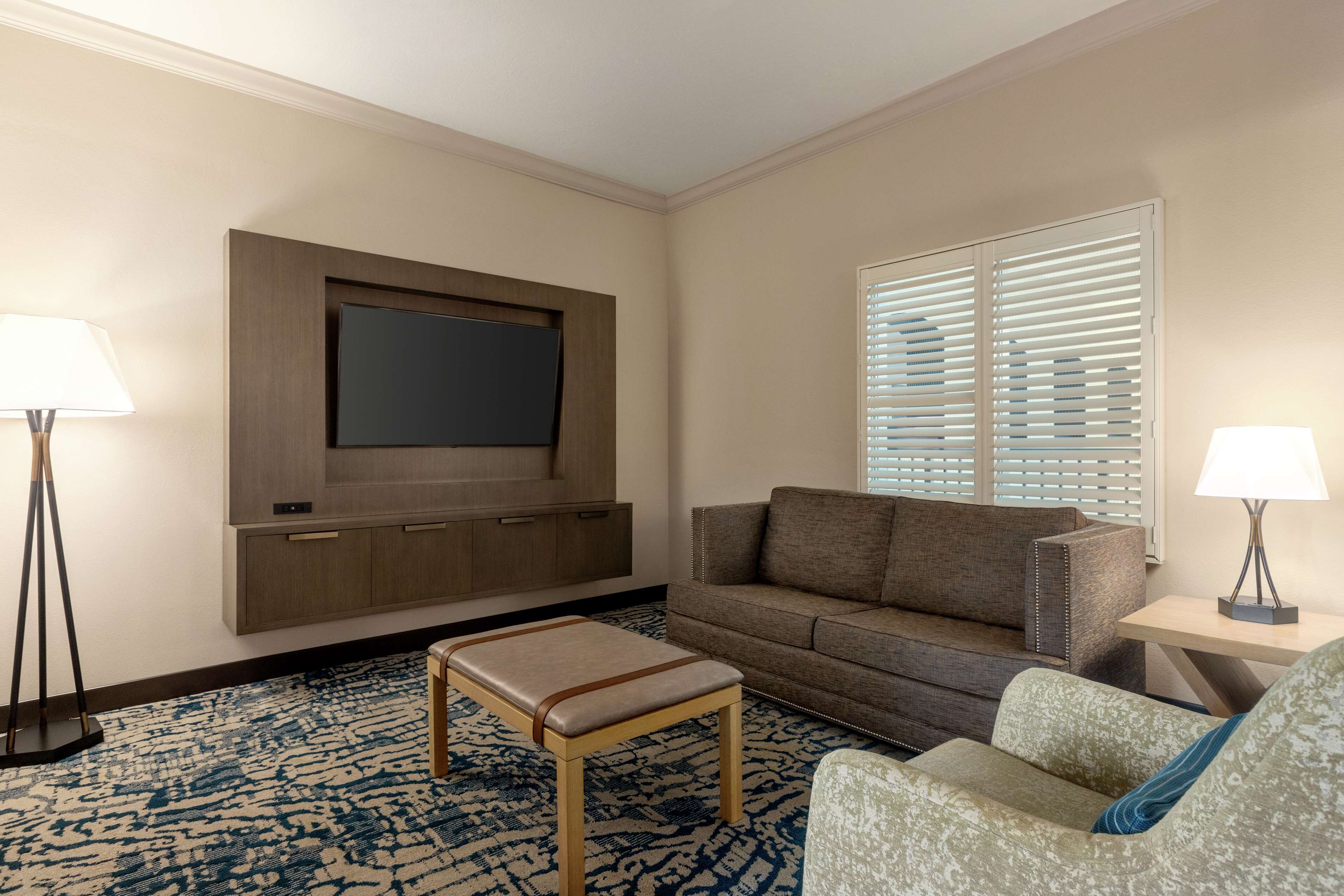 A hotel room with a flat-screen TV mounted on the wall, a sofa, a patterned rug, and two floor lamps.