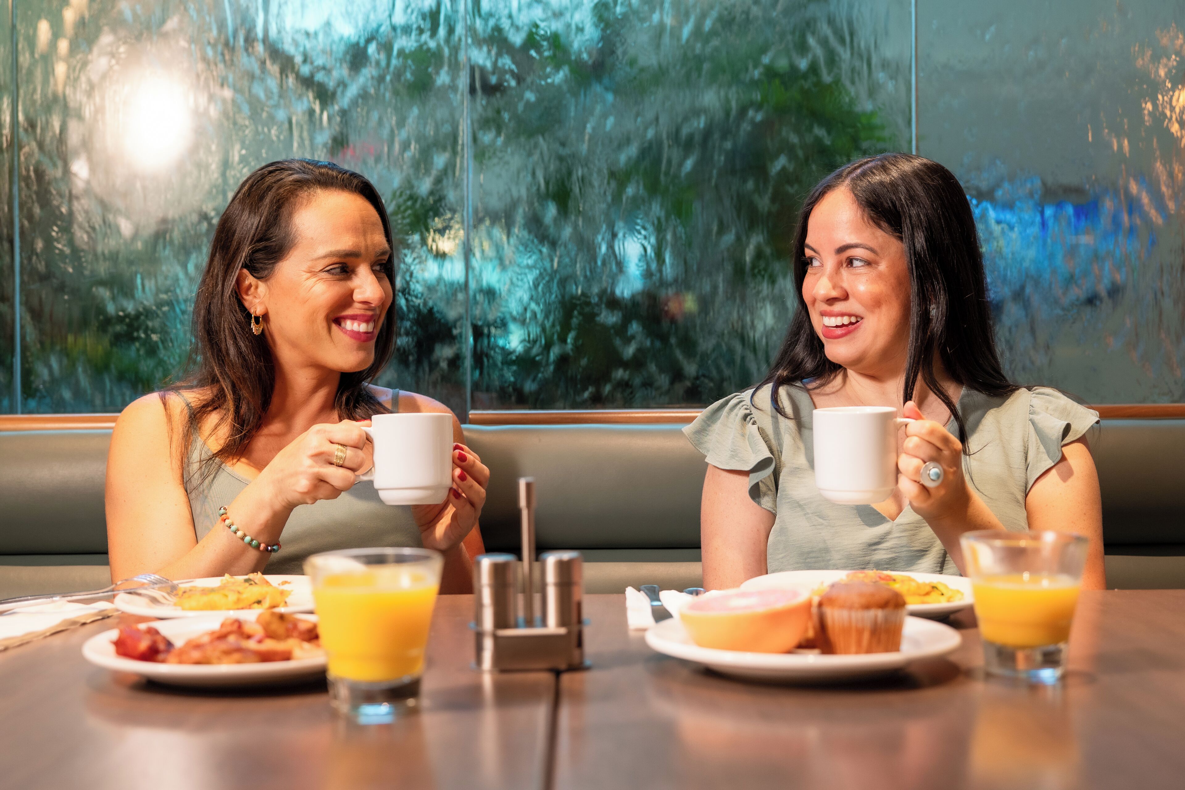 Deux personnes sont assises à une table où sont servis des aliments pour le petit-déjeuner, notamment du jus d’orange et des viennoiseries, dans une pièce d’où l’on peut voir des arbres à travers une fenêtre.