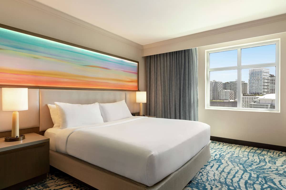 A hotel room with a large bed, two bedside lamps, a city view window, and a colorful abstract painting on the wall.