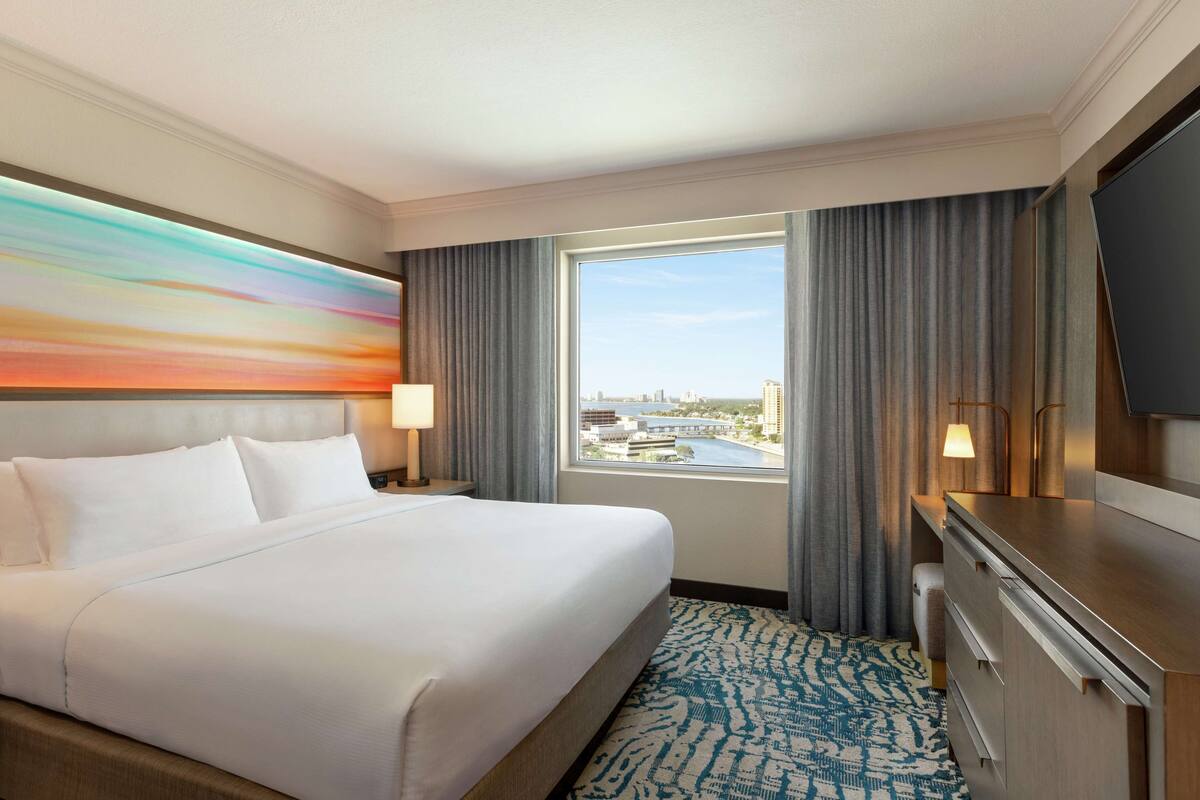 A hotel room with a large bed, a TV, a desk, and a window with a city view.