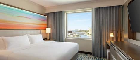 A hotel room with a large bed, a TV, a desk, and a window with a city view.