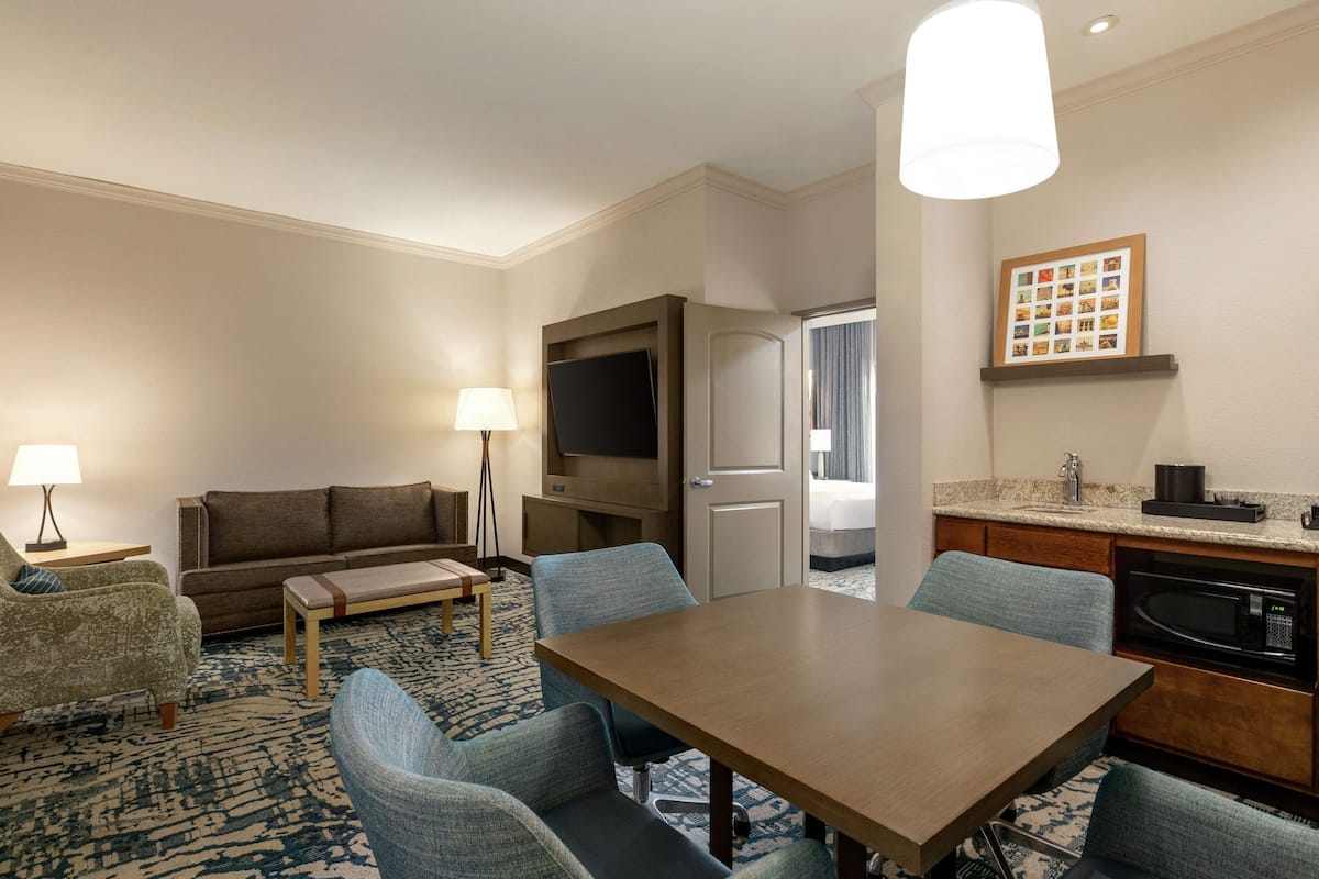 A hotel room with a dining table, chairs, a sofa, a TV, a lamp, a kitchen area with a microwave, and a framed photo on the wall.