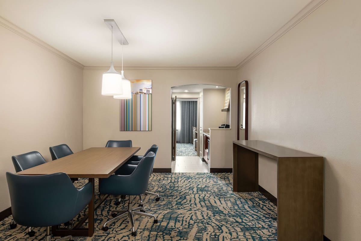 A hotel room with a wooden table, blue chairs, a patterned carpet, a hallway with a mirror, and a door leading to another room.