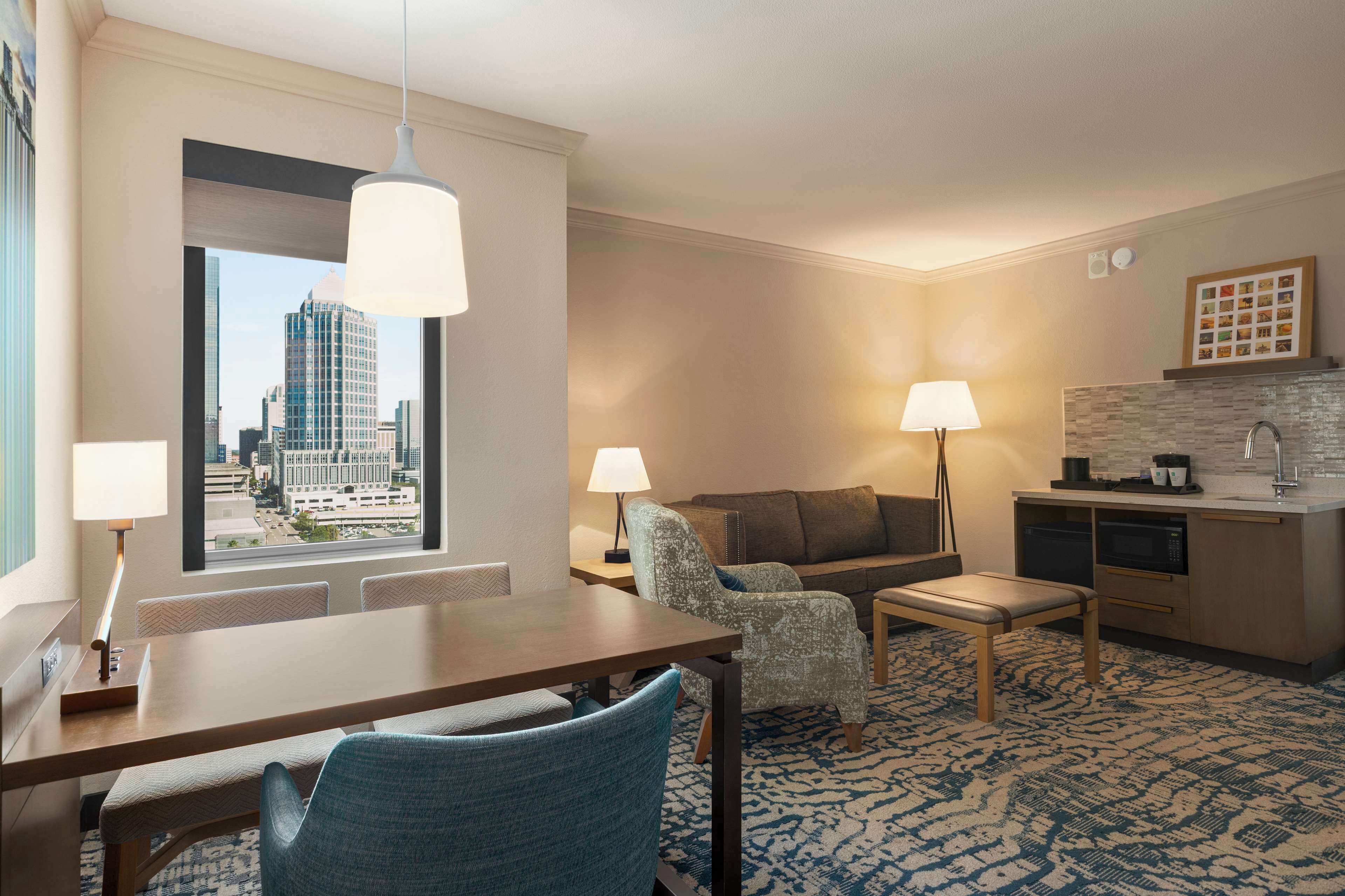 A hotel room with a dining table, two chairs, a sofa, a small coffee table, a lamp, a kitchen area, and a large window with a city view.