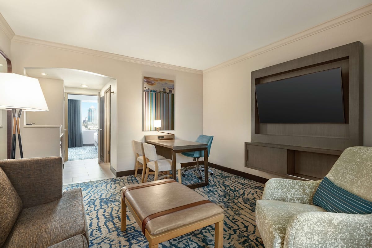 A hotel room with a seating area, a flat-screen TV mounted on the wall, a desk with a chair, and a view of the city through the window.
