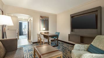 Quarto de hotel com área de estar, televisão de tela plana fixada na parede, escrivaninha com cadeira e vista para a cidade pela janela.