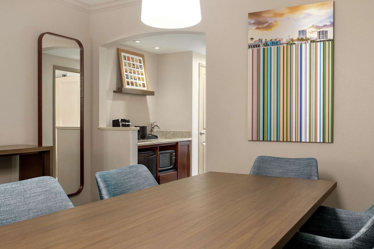 A dining area with a wooden table and blue chairs, a kitchen in the background, and a colorful striped artwork on the wall.