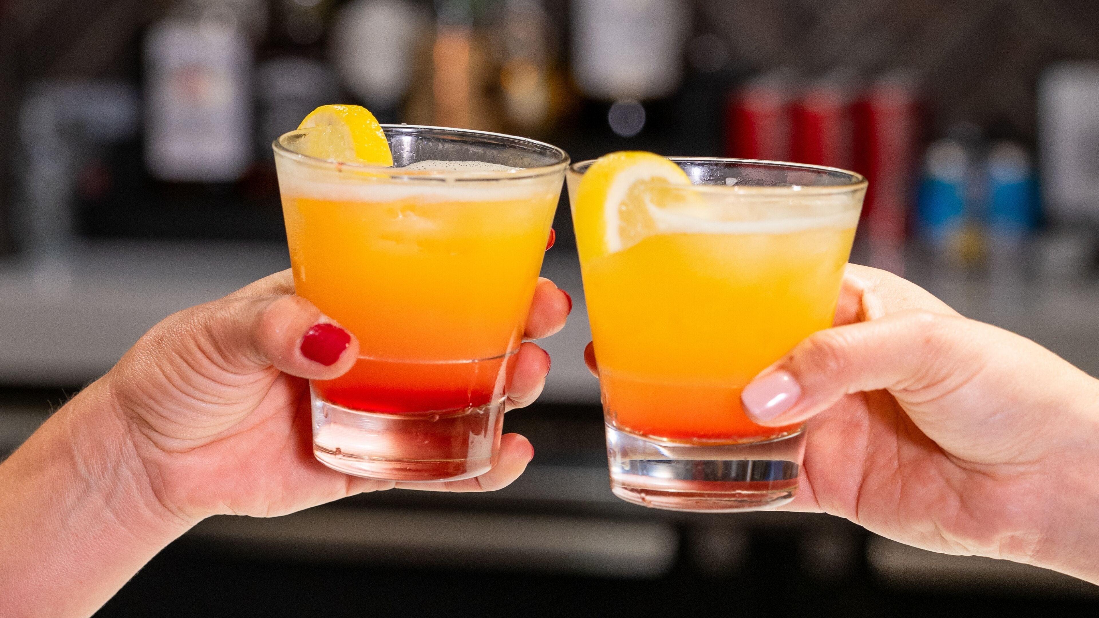 Two hands holding glasses of a layered beverage with a slice of lemon.
