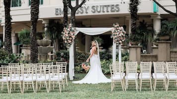 A wedding ceremony setup with a bride in a white dress, a white arch with floral decorations, and rows of white chairs.