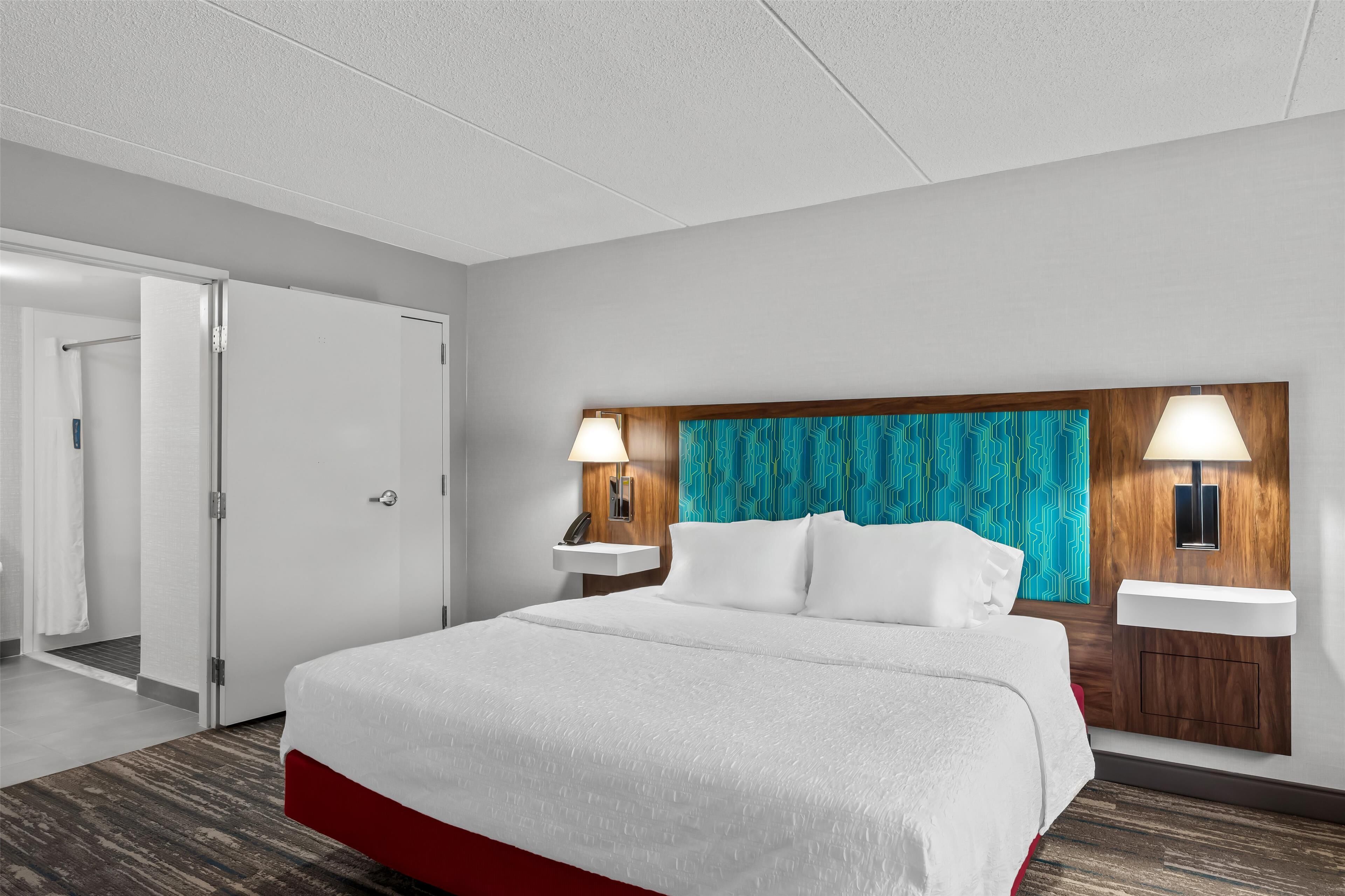 A hotel room with a large bed, two bedside tables, a headboard with a patterned curtain, and a door leading to another room.
