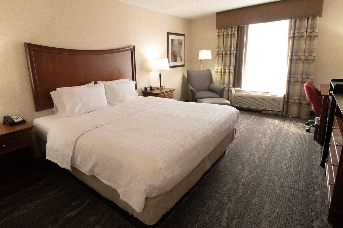 A hotel room with a large bed, a nightstand with a lamp, a chair, and a desk with a computer.