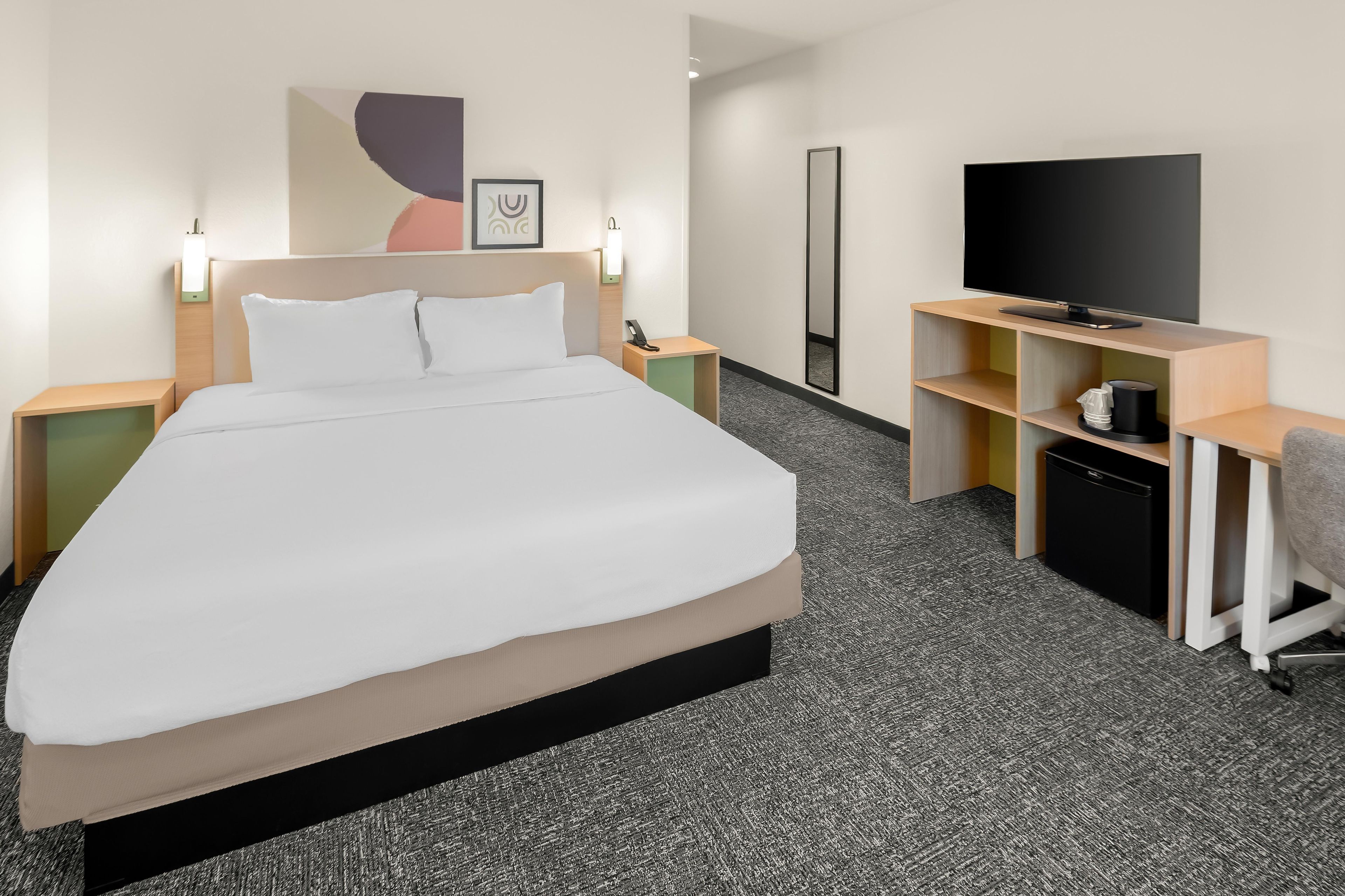 A hotel room with a bed, bedside tables, a desk with a chair, a television, and a large mirror.
