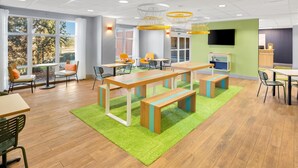 A modern indoor space with a wooden floor, green carpet, and a large table with benches. There are chairs, a TV, and a window with a view of trees. - Spark by Hilton Grand Island (Grand Island)