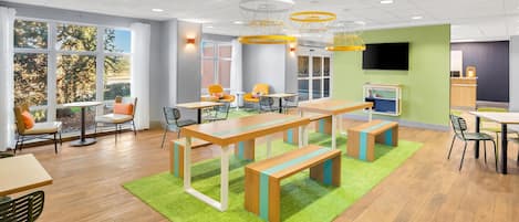 Un espace intérieur moderne doté d’un plancher en bois, d’une moquette verte et d’une grande table avec des bancs. On y trouve également des chaises, un téléviseur et une fenêtre donnant sur des arbres.