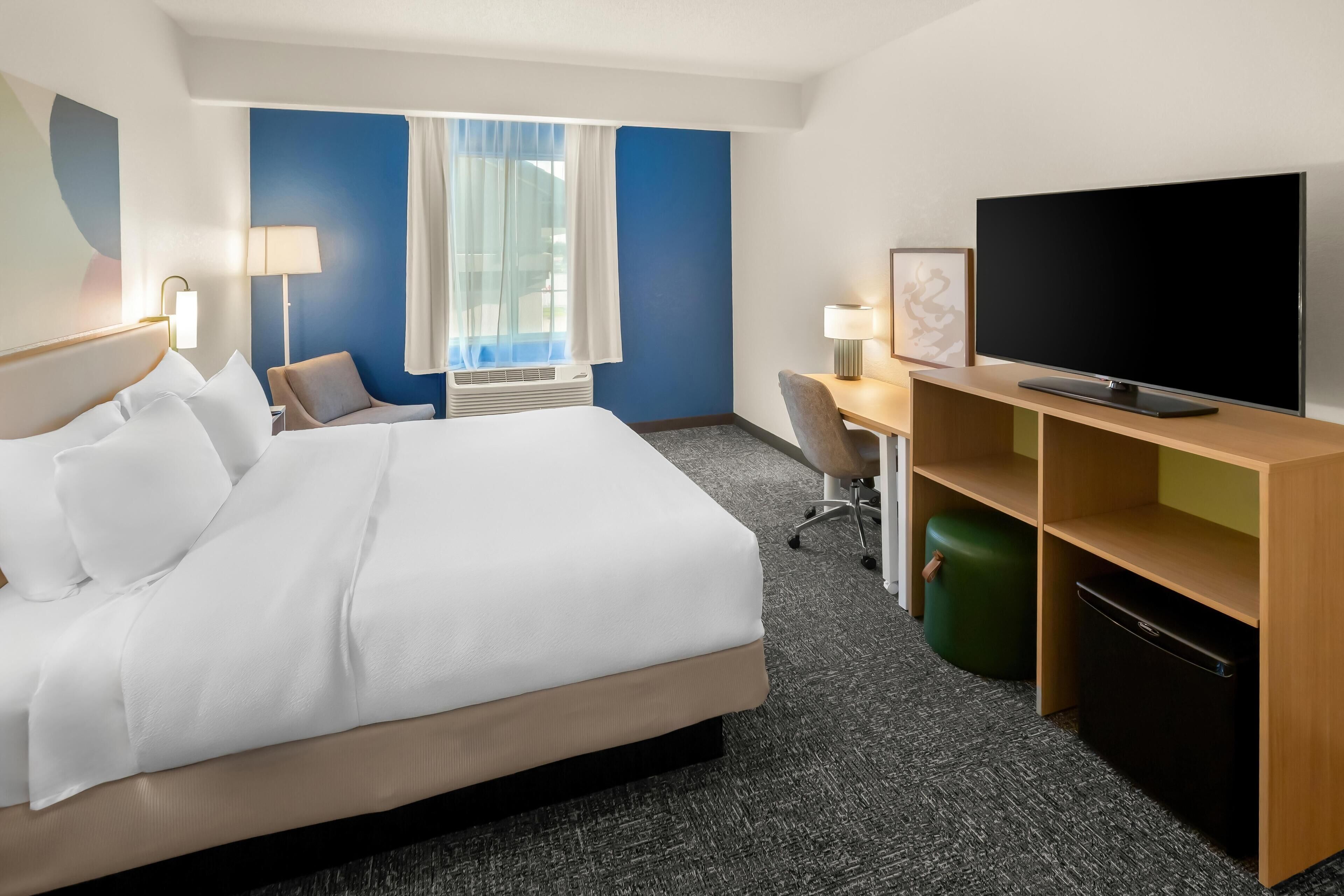 A hotel room with a bed, desk, chair, television, and a small green trash bin.