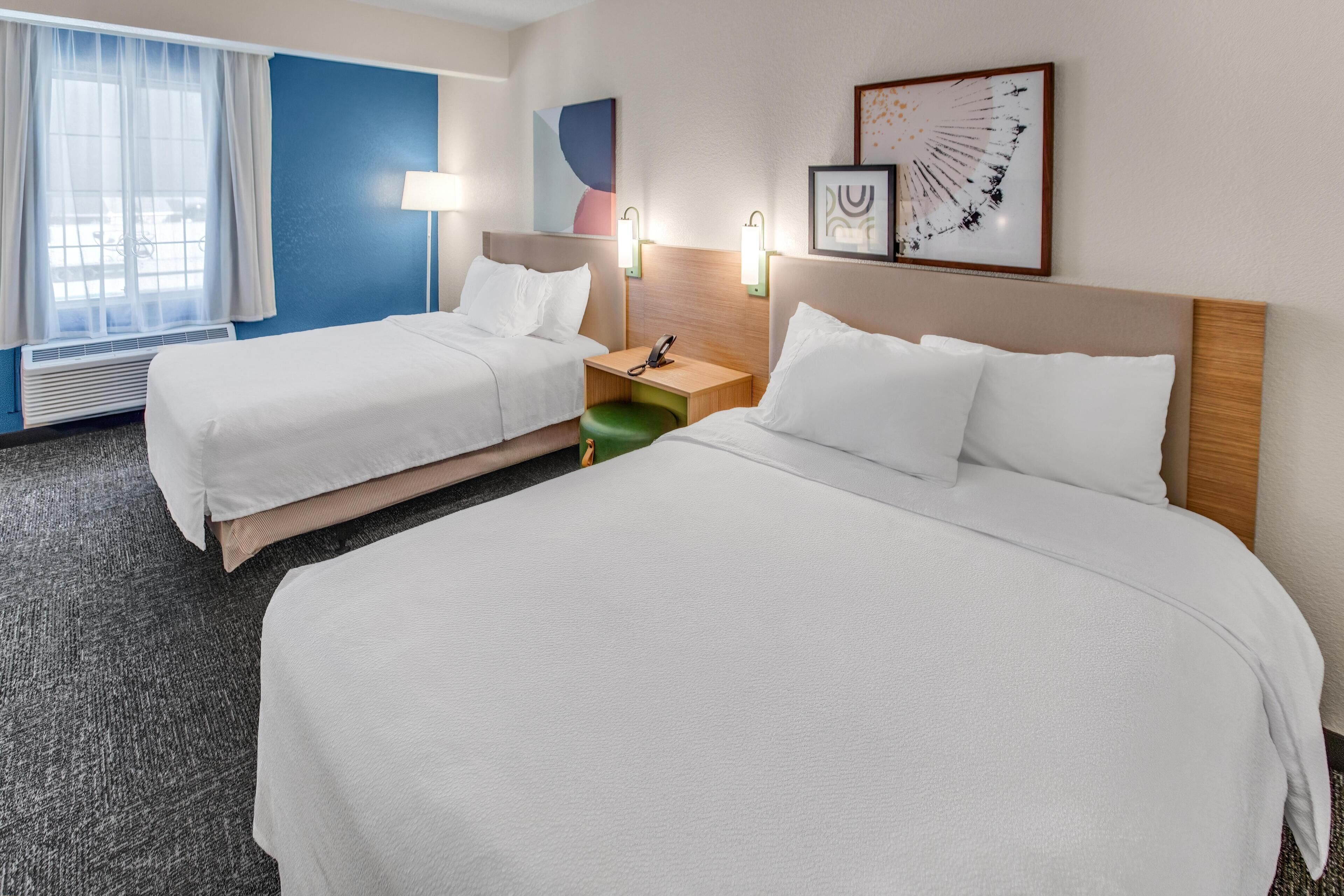 A hotel room with two beds, a window with curtains, a wall-mounted lamp, and artwork.