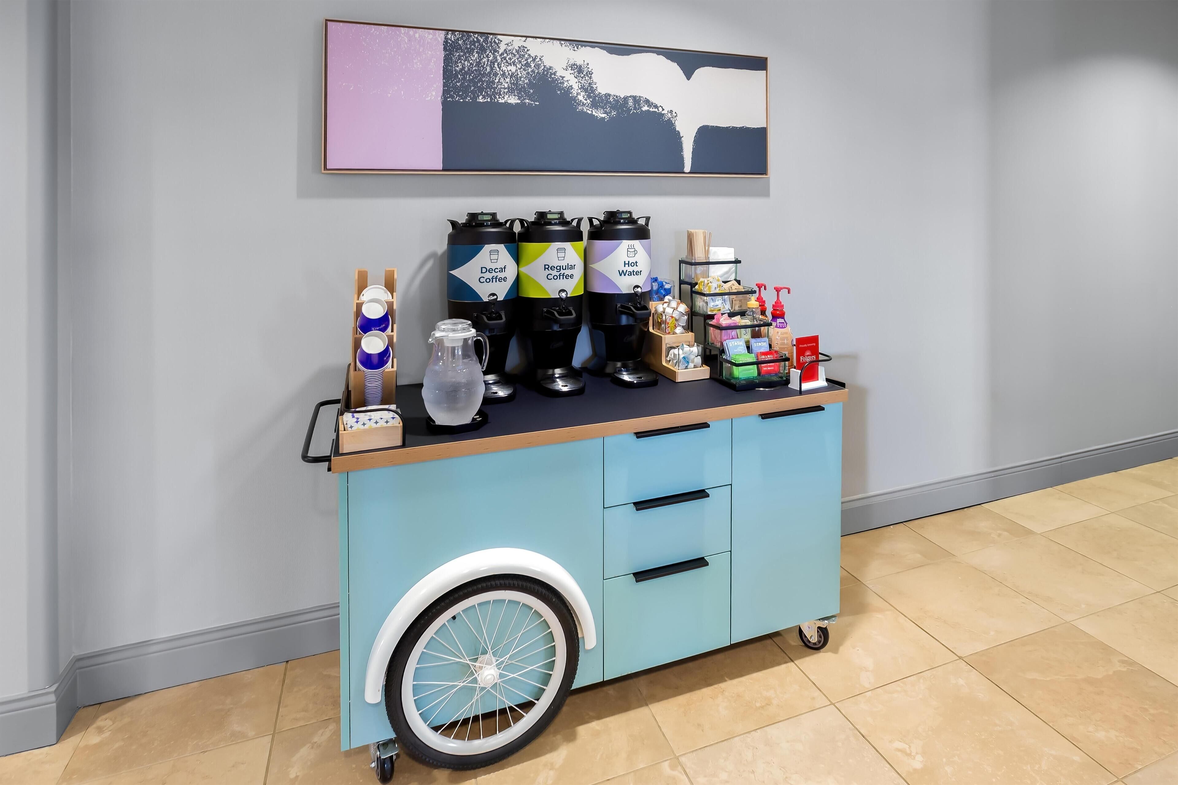 Uma estação de café à disposição dos hóspedes, com dispensadores de café descafeinado, café normal e água quente, um jarro e uma seleção de xícaras de café da marca.