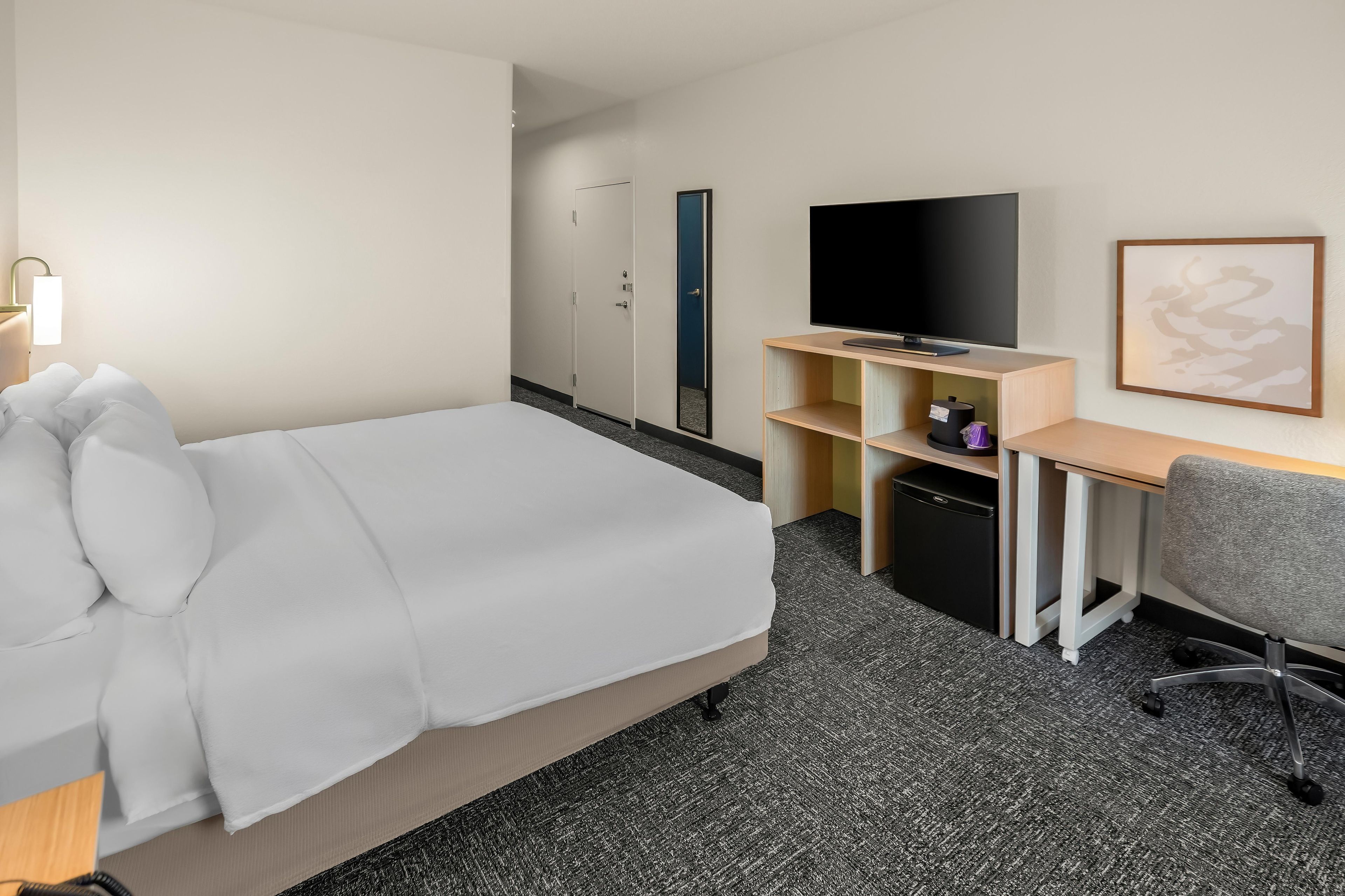 Habitación de hotel con cama, escritorio, televisión, silla y un refrigerador pequeño.