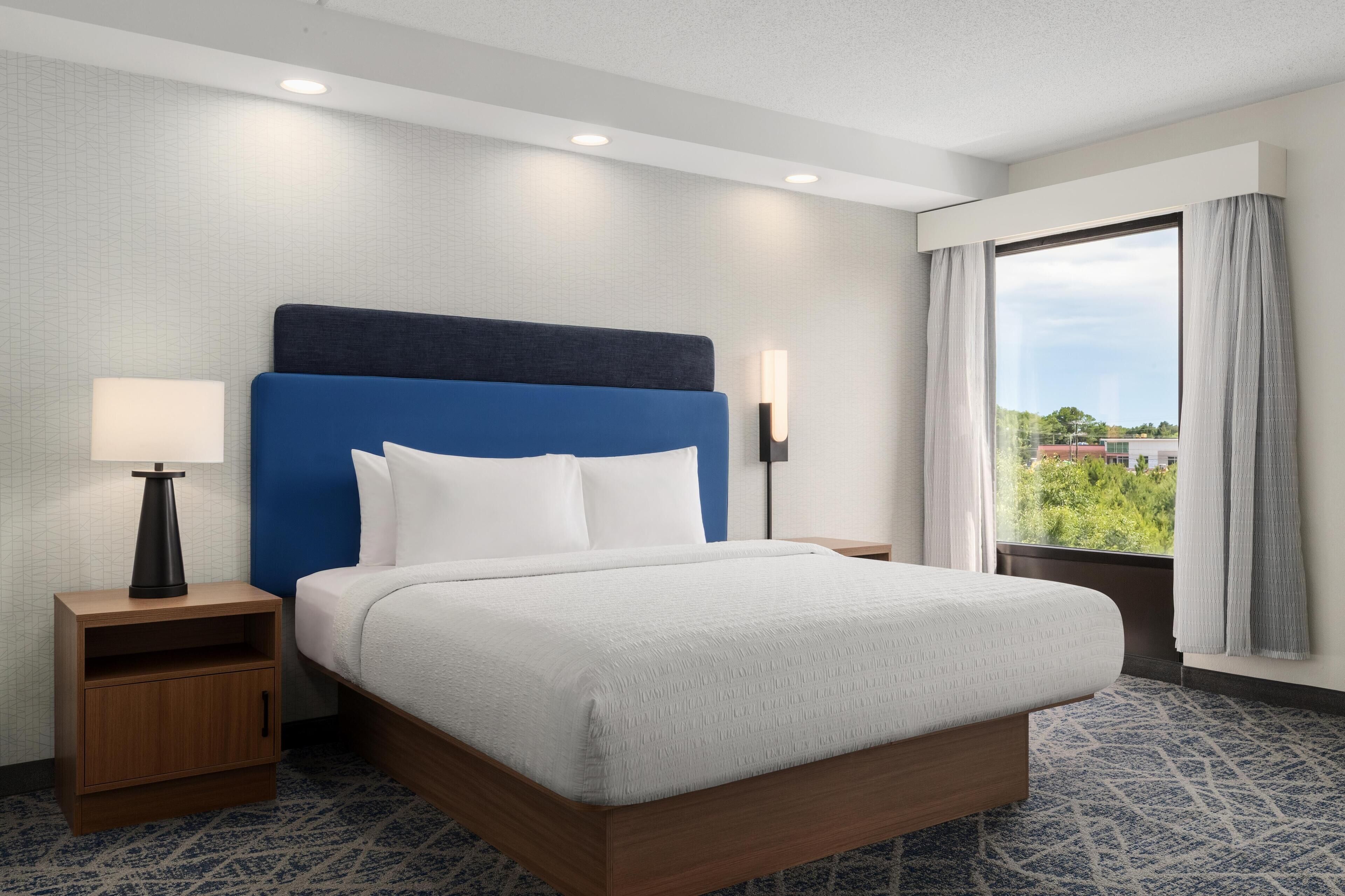 A hotel room with a bed, a nightstand, a lamp, a window with curtains, and a view of greenery outside.