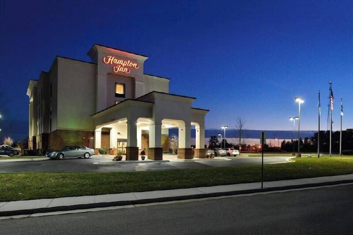 Ein Hampton Inn Hotel mit moderner Architektur, das ein auffälliges Neon-Schild, einen gut beleuchteten Eingang und einen Parkplatz zeigt.