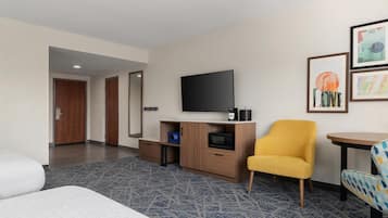 A hotel room with a bed, a television, a small desk with a microwave, a yellow armchair, and a framed picture on the wall.