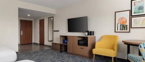 Una camera d'albergo con un letto, una televisione, una piccola scrivania con un forno a microonde, una poltrona gialla e un quadro appeso al muro.