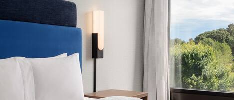 Quarto de hotel com uma cama, uma mesa de cabeceira, uma luminária na parede e uma janela com vista para o verde.