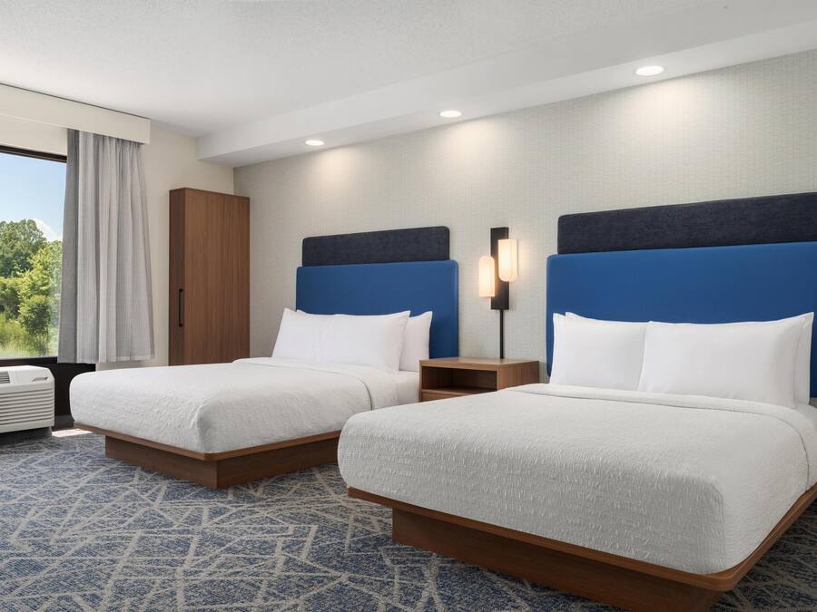 A hotel room with two beds, a wardrobe, a nightstand, and a window with a view of greenery.