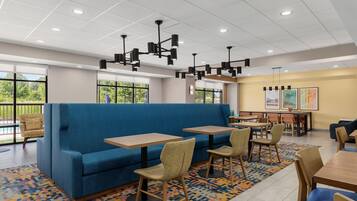 Un interno ristorante moderno con un divano blu, tavoli e sedie in legno, e ampie finestre che si affacciano sul verde.