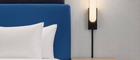 A bed with a blue headboard, white pillows, and a white blanket. There is a bedside table with a wooden finish and a wall-mounted lamp.