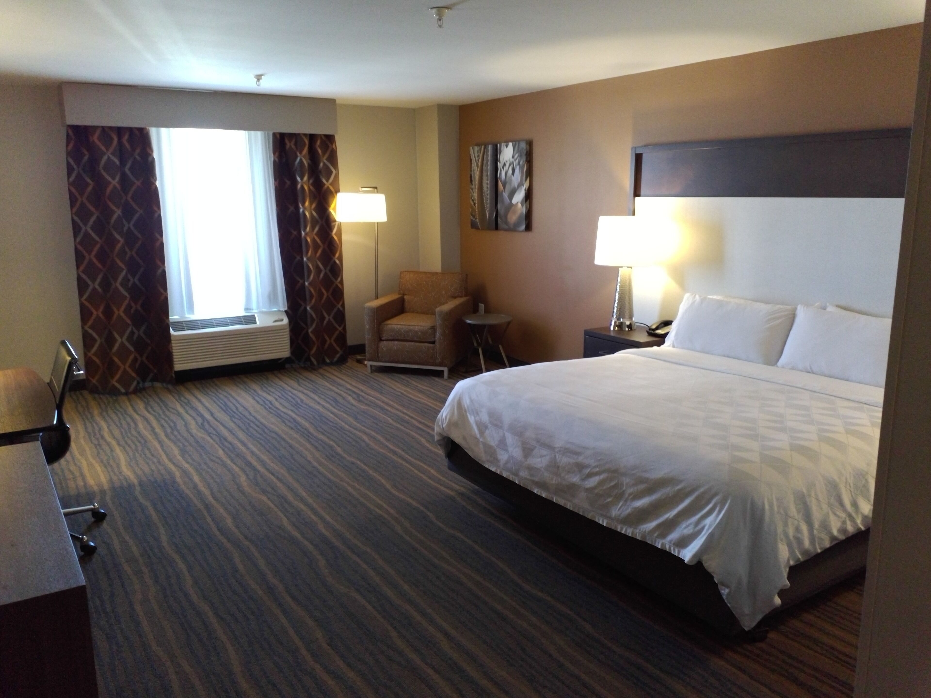 A hotel room with a large bed, a chair, a desk, a lamp, and a painting on the wall.