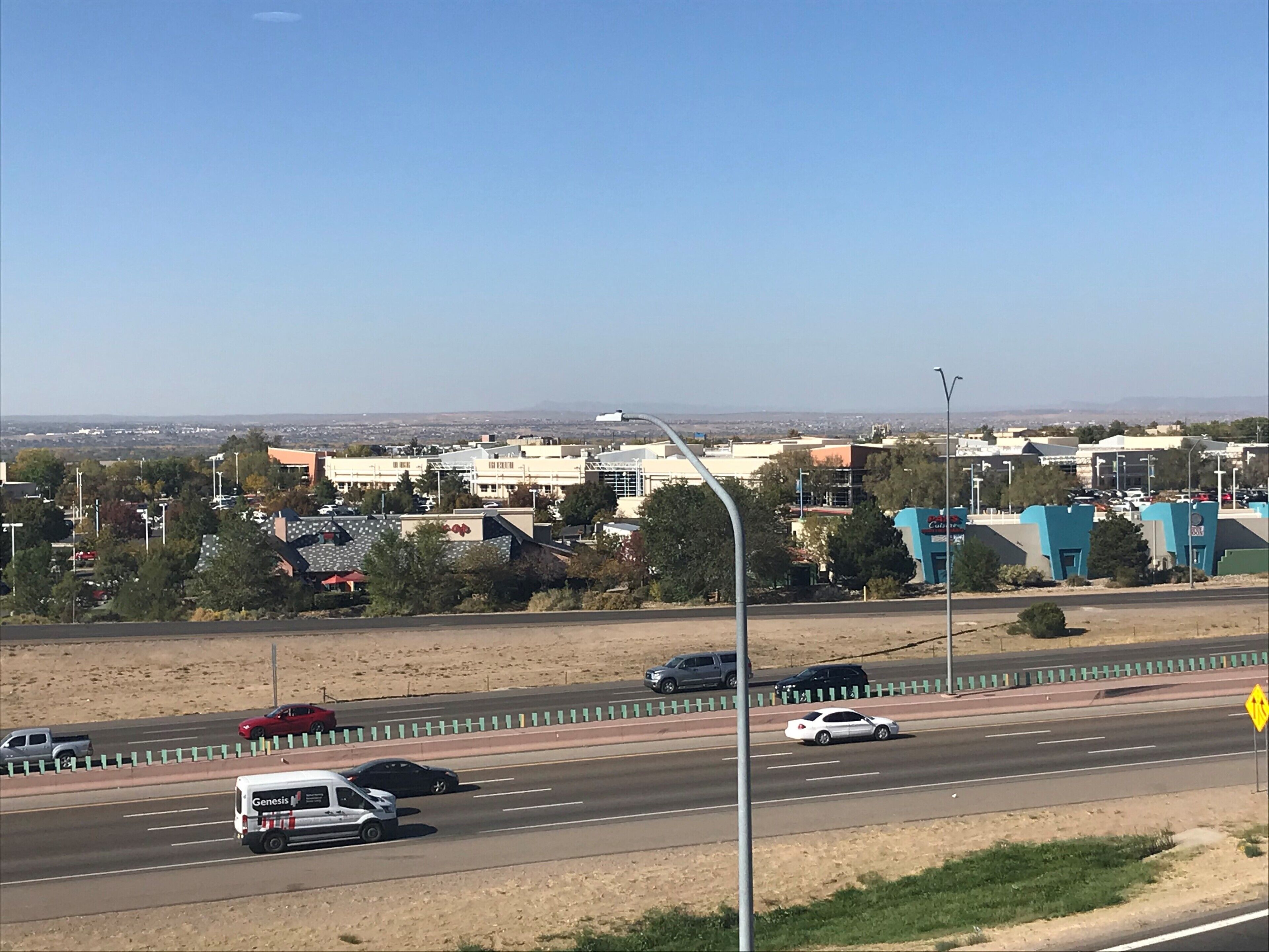 A highway with multiple lanes and vehicles, including cars and a van. There are buildings and trees in the background.