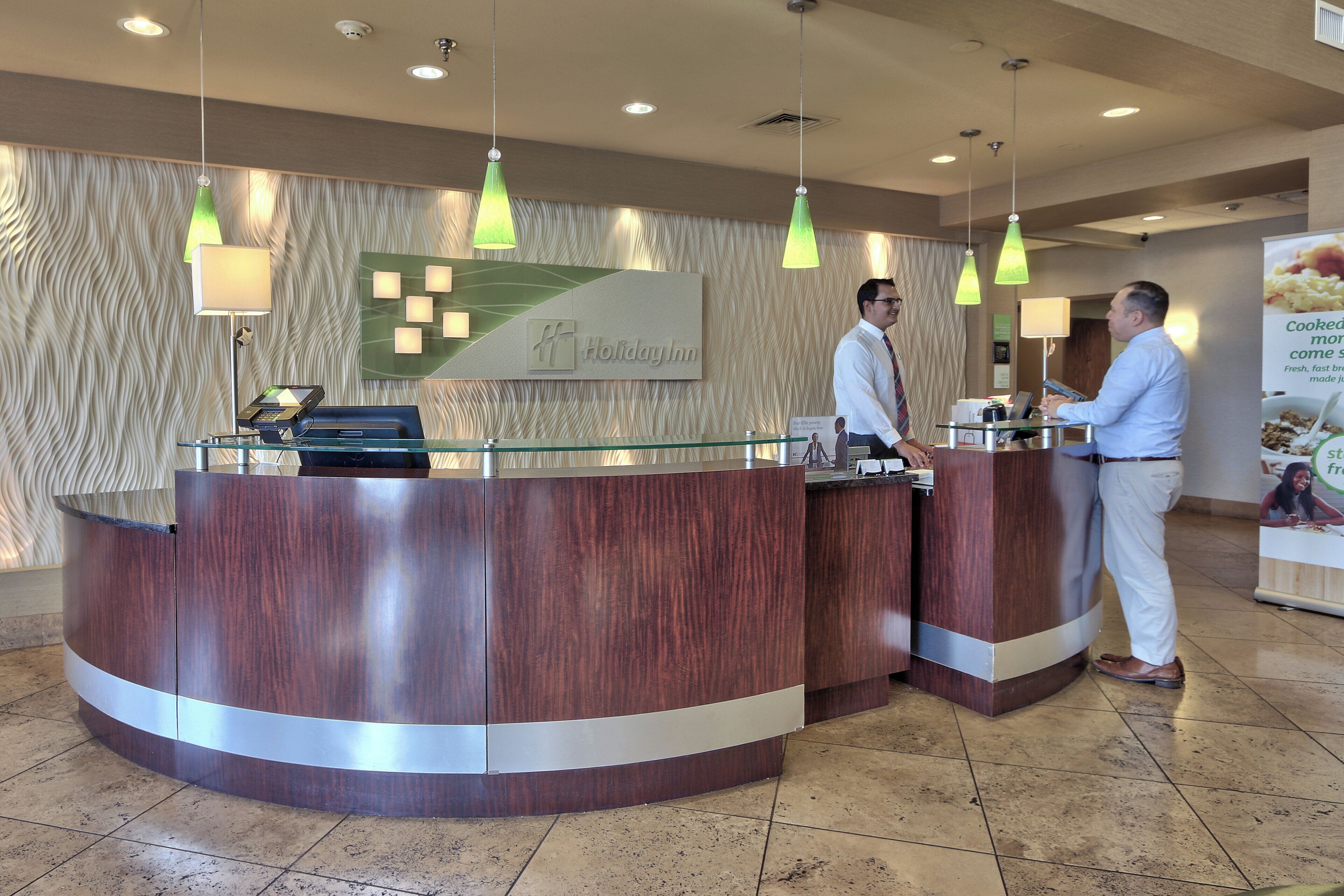 A reception desk with a wooden finish, a digital check-in kiosk, and a sign for 'Holiday Inn'.