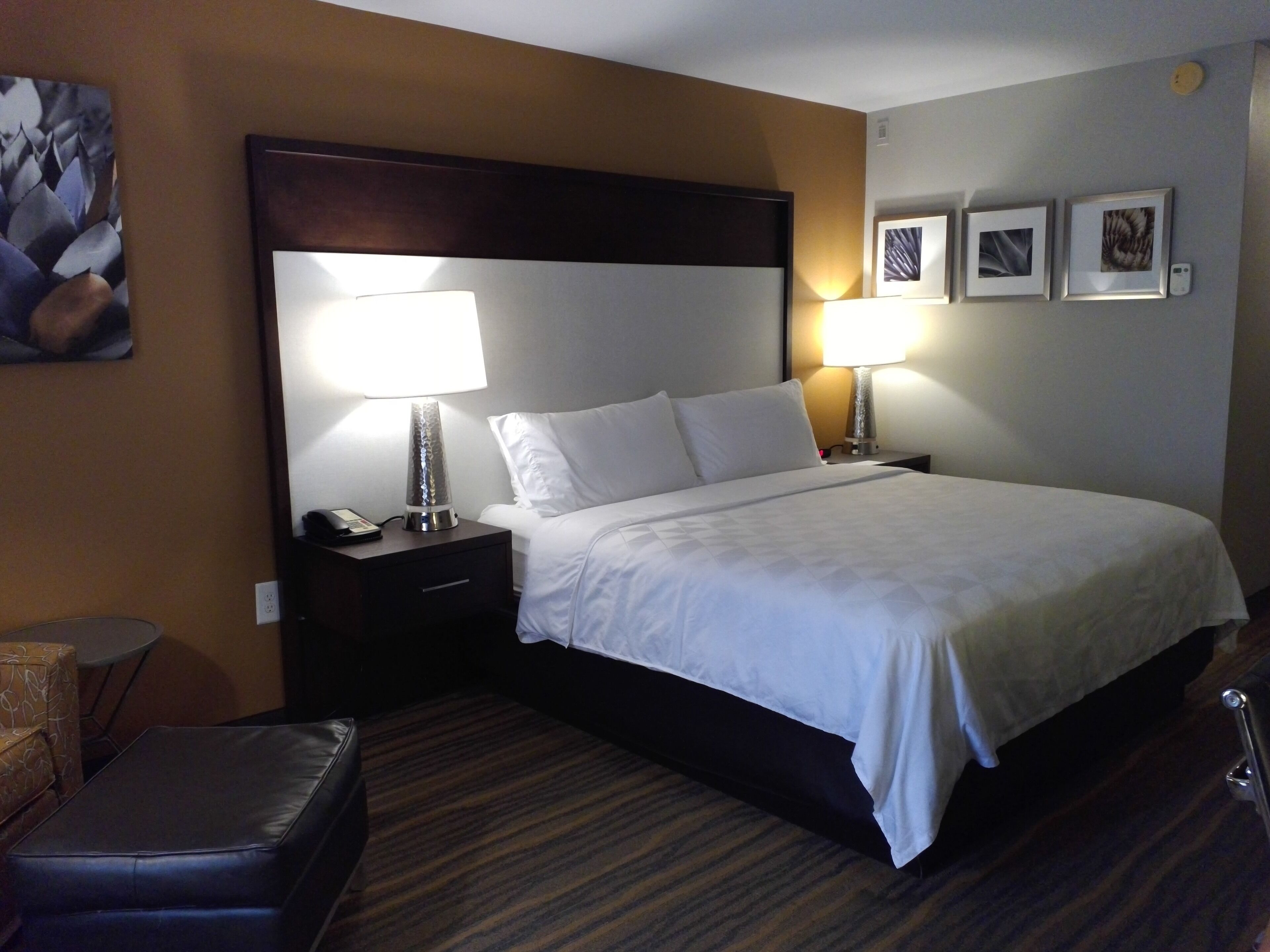 A hotel room with a large bed, two bedside lamps, a nightstand with a phone, a small table with a chair, and a wall-mounted artwork.