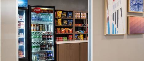A vending machine stocked with beverages and snacks, a refrigerator with drinks, and shelves with packaged food items.