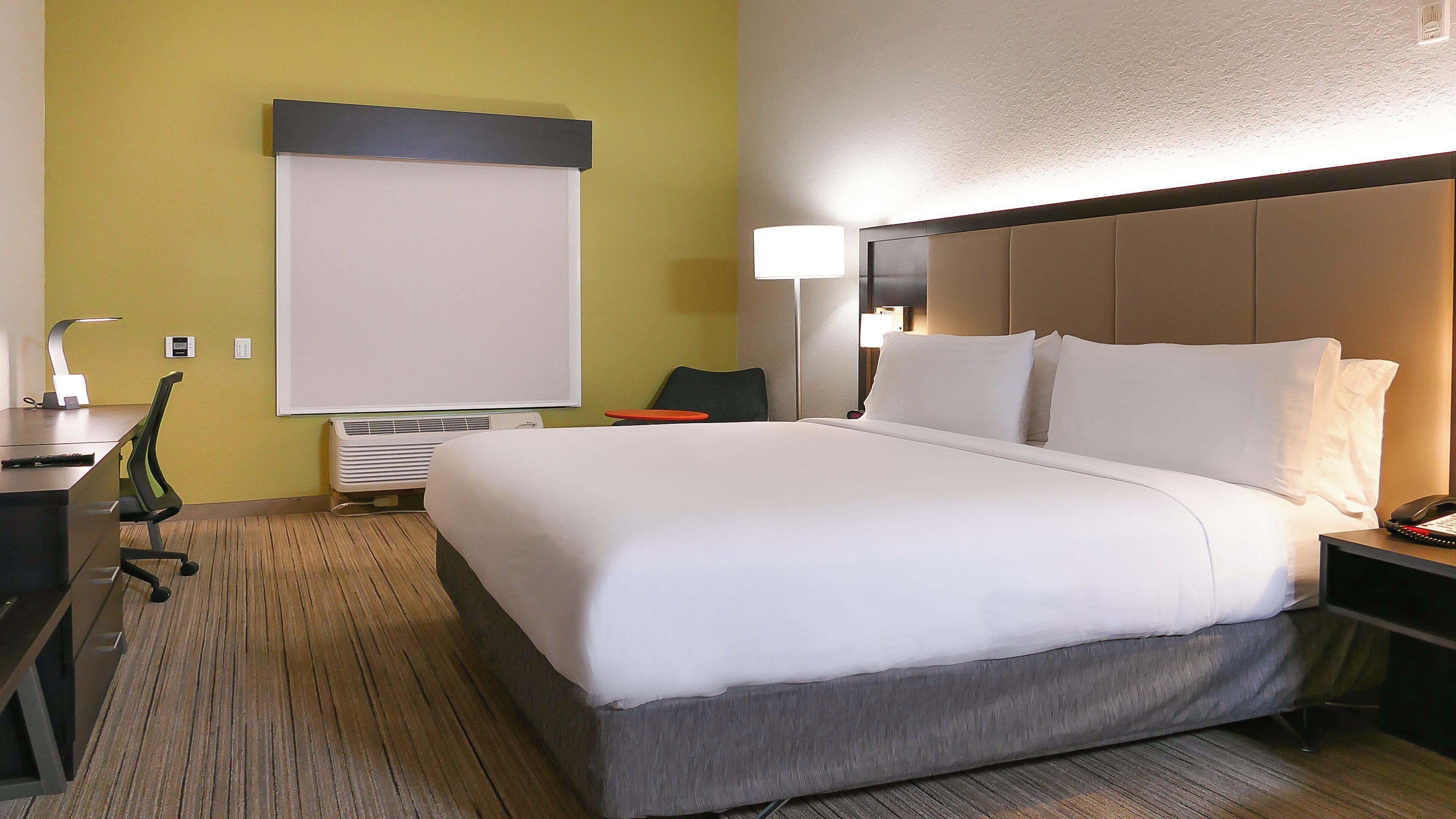 Une chambre d’hôtel avec un grand lit, un bureau avec une chaise, une table de chevet avec une lampe et un climatiseur fixé au mur.