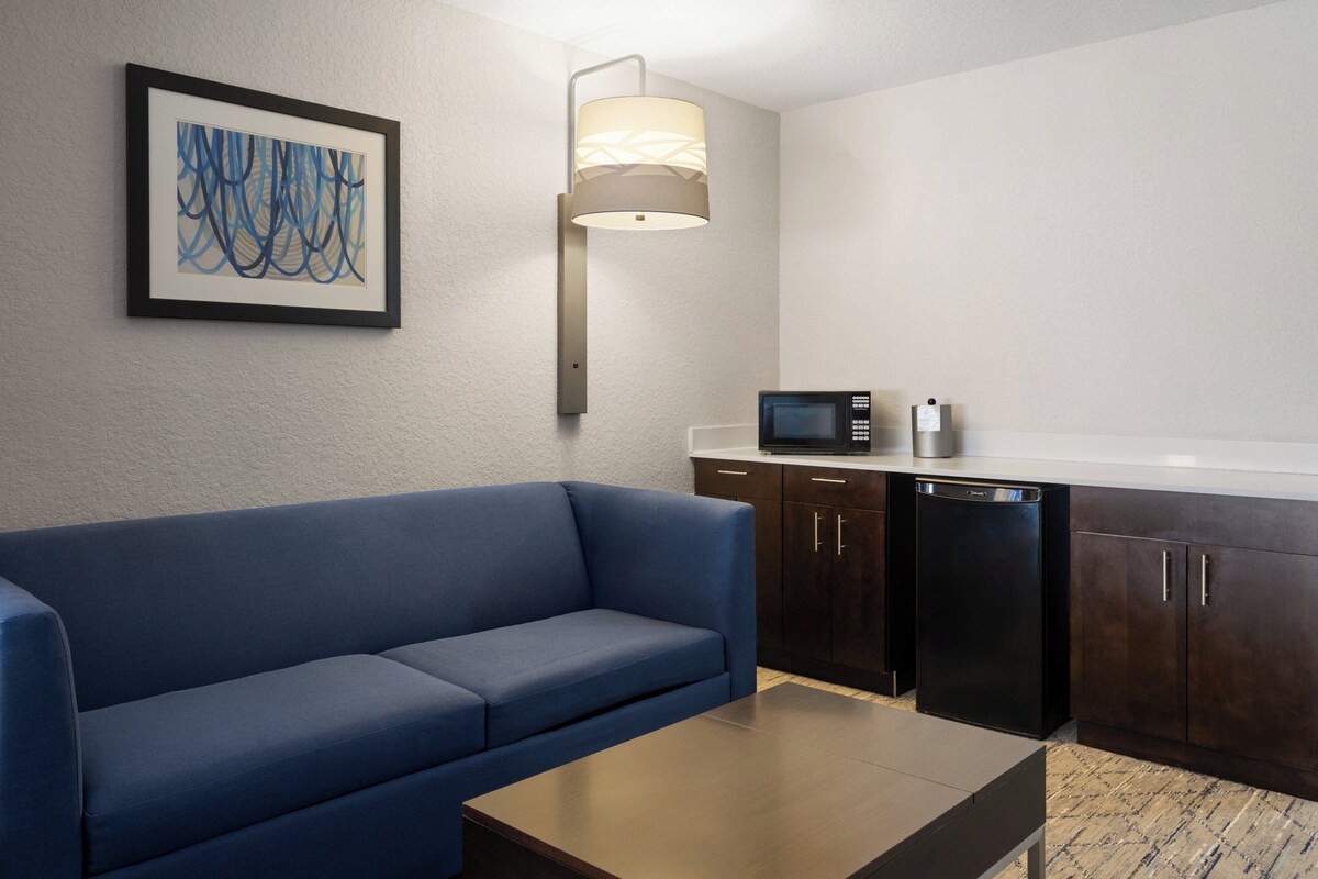 Quarto de hotel moderno com sofá azul, uma pequena mesa, luminária de parede, obra de arte abstrata emoldurada, micro-ondas e armários de madeira escura.