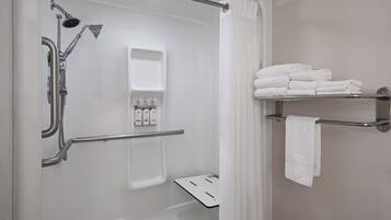 Un espacio de ducha moderno con ducha de mano, ducha de lluvia y asiento integrado. Hay toallas blancas apiladas en un estante y una toalla colgada en un perchero.