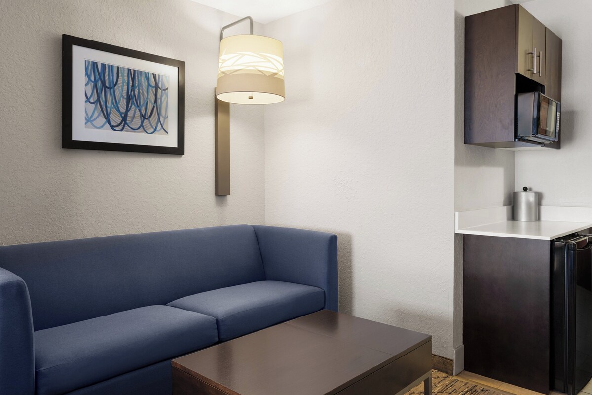 Quarto de hotel moderno com um sofá azul, uma mesinha, uma luminária de parede, um quadro abstrato emoldurado e uma kitchenette com micro-ondas e chaleira.